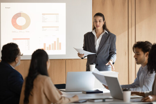 Educational presentation. Corporate learning session lead by young woman professional coach teacher trainer presenting financial data project insights to diverse multicultural team on screen and paper