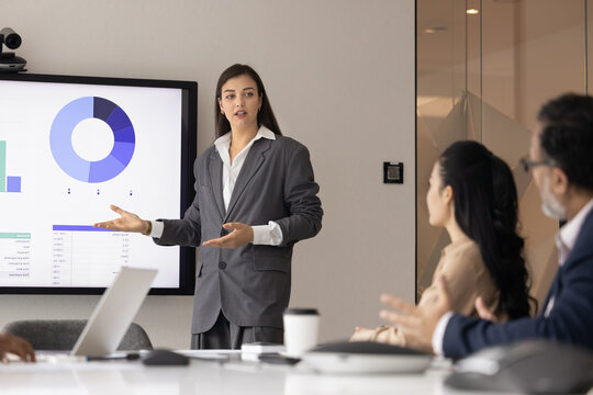 Pitch meeting. Young businesswoman stand by boardroom table speak to group of multicultural colleagues lead presentation focused on launching new project offer marketing strategy stats data on screen