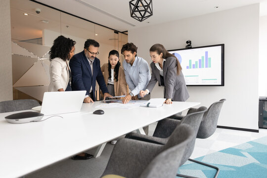 Project planning. Professional multiethnic business team ahead with mature man leader stand around table in conference room have creative brainstorming session discuss ideas use papers digital devices