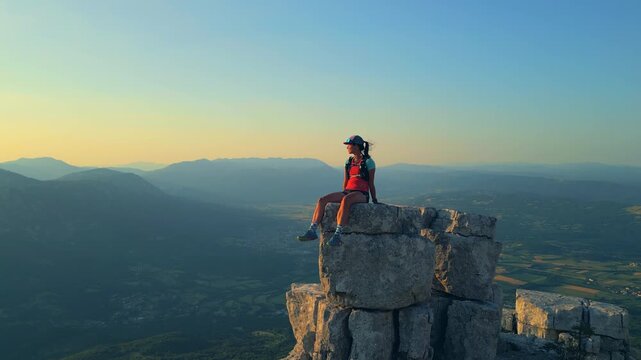 A determined female runner triumphantly sits atop a rugged mountain peak, capturing achievement and joy. The breathtaking landscape below highlights endurance and adventure.
