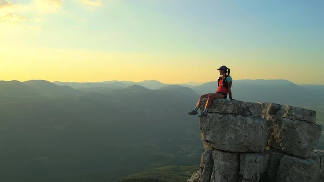 A determined female runner triumphantly sits atop a rugged cliff, overlooking expansive mountain ranges. Golden hour light highlights her achievement and endurance.