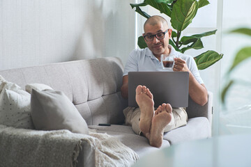 Mature freelancer working from home, enjoying coffee and using laptop on sofa