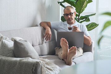 Mature freelancer working from home, enjoying coffee and using laptop on sofa