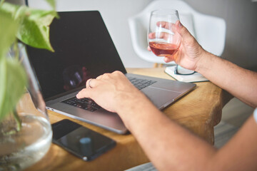 Freelancer enjoying a glass of wine while working from home on his laptop, combining work and relaxation