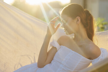 A young woman in a white dress enjoying a peaceful moment in a hammock under soft sunlight, evoking...