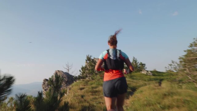 A female runner in athletic gear and a hydration pack sprints through a scenic grassy trail with rocky outcrops and pine trees under a clear blue sky, evoking endurance, achievement.