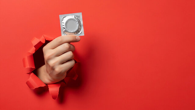 A hand emerges from a torn red background, holding a condom. This impactful image promotes safe sex, contraception, and sexual health awareness with a dynamic, modern aesthetic.

