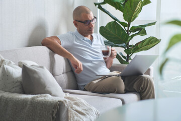 Man working from home, enjoying a cup of coffee while using his laptop on the sofa