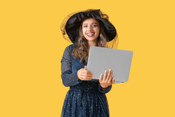 Young woman dressed as witch for Halloween with laptop shopping online on yellow background