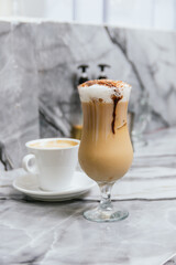 A glass of coffee with a chocolate topping sits on a table next to a white cup