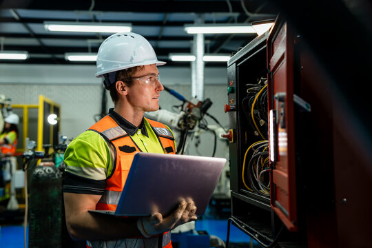 A man in a yellow and green vest is looking at a laptop computer. Electrician or technician working on the wiring and diagnostics of an automated robotics system in a factory.