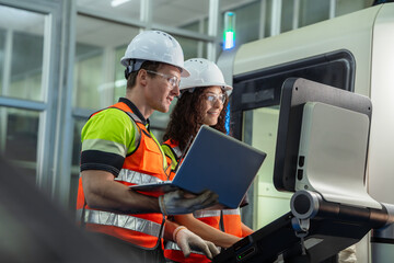 Two people wearing safety gear and holding a laptop. Team of diverse engineers, male and female, programming and operating a CNC machine in a modern factory.