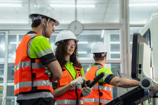 Three people wearing safety gear and orange vests. Group of machinists in an on-the-job training session, learning to program and control an automated industrial machine.