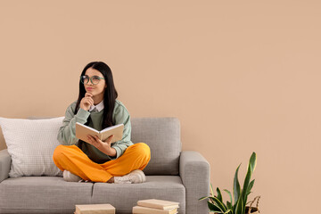 Thoughtful young woman reading book on sofa near beige wall