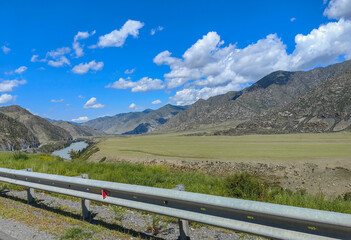 The panoramic view showcases mountains and a river under a vibrant blue sky with fluffy clouds.