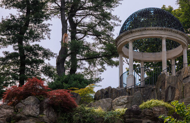 Classical stone pavilion with a domed roof set among trees and rocks in a landscaped garden. A historic architectural structure symbolizing culture, heritage, and timeless outdoor design.