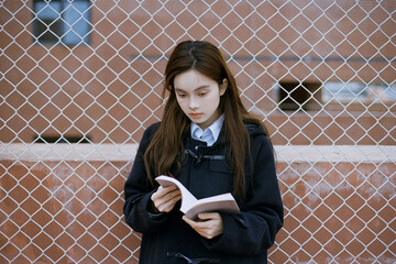 Young female reading book near chain-link fence.