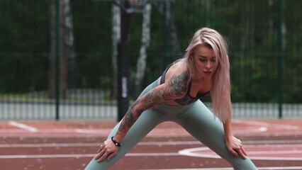 Young blonde woman in sportswear doing stretching exercises standing on basketball court in the morning. Woman feels pleasantly tired after intense daily workout program. Genuine candid photo
