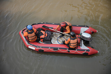 Rescuers in orange uniforms and helmets row a red rubber boat in murky water during a search and...