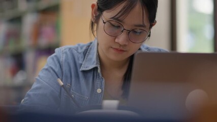Young Asian woman, early 20s, wearing glasses and casual clothes, studying in a library using a laptop and book. Ideal for concepts like education, learning, focus, university, or academic life. - Powered by Adobe