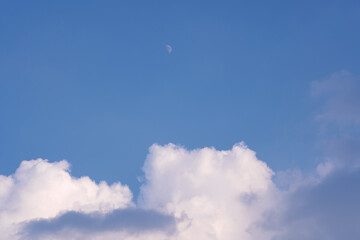 Dramatic sky with blue tones, fluffy white clouds, dark storm clouds, and a visible daytime moon. Peaceful yet moody natural sky background.
