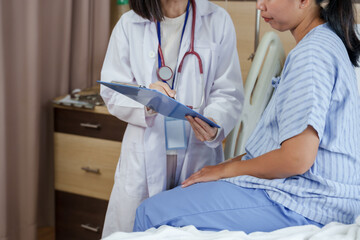 Asian female doctor holding clipboard discussing treatment details with female patient sitting on hospital bed during medical consultation inside ward