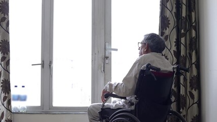 Elderly indian man on wheelchair feel lonely in house looking out of window. Retired disable male lost in thoughts. Senior person Solitude concept. 4k