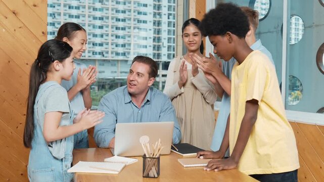 Skilled teacher clapping hand to encourage student to do project or celebrating successful project. Diverse children clap hands together while standing surround professional instructor. Edification