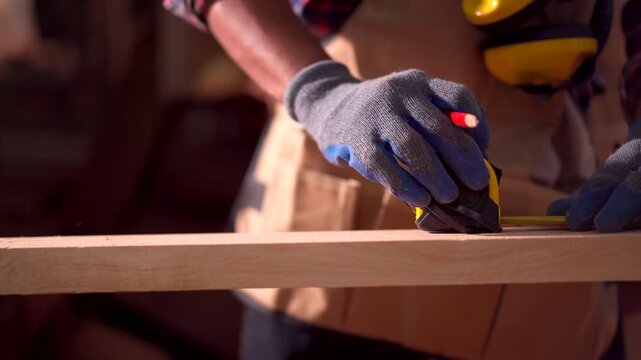 Carpenter works in a woodworking shop, using a tape measuring and pencil to mark on a wood plank. 