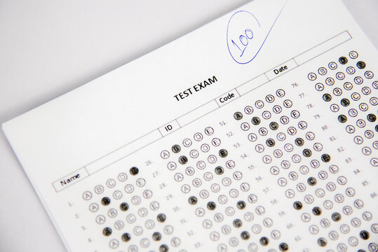 shows a hand holding a blue pen and writing the number "100" inside a circle on a test paper. The paper appears to be a completed multiple choice answer sheet, with some of the circles filled in. 