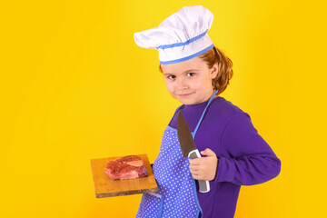 Child cook hold cutting board with meat beef steak and knife. Portrait of little child in uniform of cook. Chef boy isolated on studio background. Cute child to be a chef. Child dressed as a chef hat.