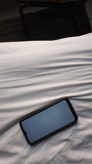 Top view of a mobile phone with a blank white screen on white bedsheet. Technology mockup template.