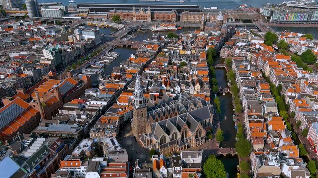 4K drone view of Amsterdam&rsquo;s historic core : the Gothic Oude Kerk ringed by canals and gabled houses, with Amsterdam Central Station and the IJ waterfront in the distance.