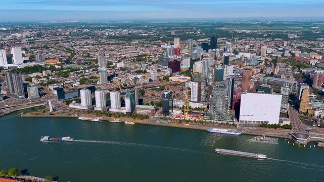 4K drone panorama of Rotterdam&rsquo;s skyline along the Nieuwe Maas, showcasing modern high-rises, waterfront promenades and cargo barges moving through the busy Dutch port city.
