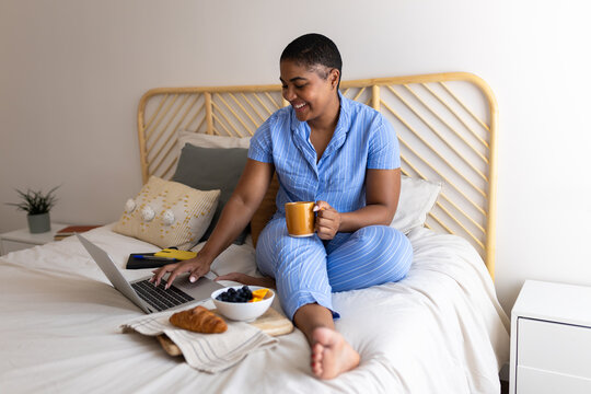 Smiling Black woman using laptop and having breakfast in bed in pajamas