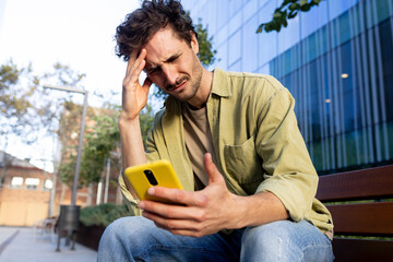 Frustrated young man reading bad news on mobile phone outdoors.