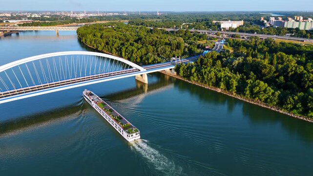 Long riverboat travelling by the Danube in Bratislava, Slovakia. Water vehicle goes under the Appolo Bridge. Aerial view on the lush greenery at the waterfront.