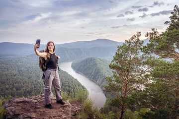 A hiker stands atop a rocky ledge, capturing a selfie against a breathtaking backdrop of lush green...