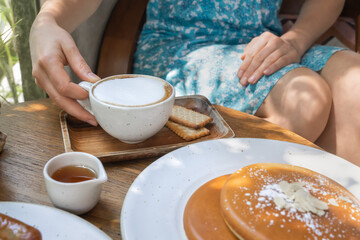 Brunch with pancakes in the morning in a sunny summer cafe. A woman is drinking coffee, and there are fragrant pancakes and maple syrup on a wooden table.