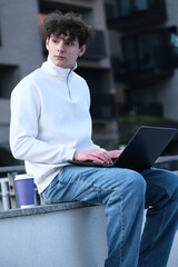 young male student with coffee to go sitting in park and using laptop while working on assignment