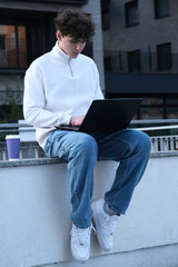 young male student with coffee to go sitting in park and using laptop while working on assignment