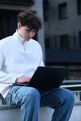 young male student with coffee to go sitting in park and using laptop while working on assignment