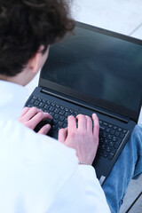 young male student with coffee to go sitting in park and using laptop while working on assignment