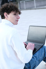 young male student with coffee to go sitting in park and using laptop while working on assignment