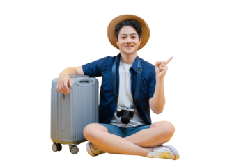 A young man ready for his next travel adventure, sitting next to his luggage. He points to the right while smiling, wearing a travel hat with a camera, ready to enjoy his trip