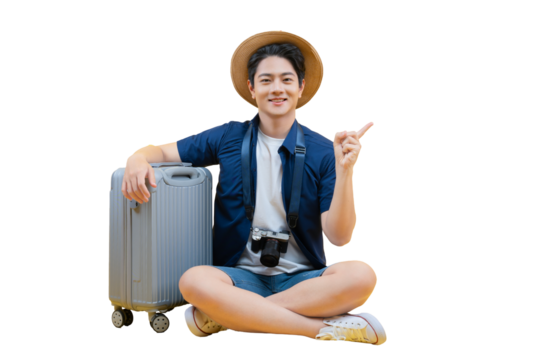 A young man ready for his next travel adventure, sitting next to his luggage. He points to the right while smiling, wearing a travel hat with a camera, ready to enjoy his trip