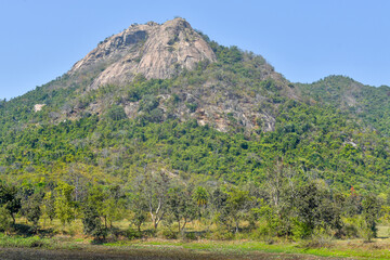 A rugged hill rises prominently against a clear blue sky, its rocky summit crowned by large granite...