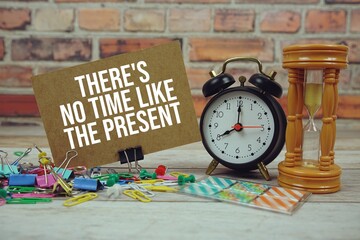 There's no Time Like the Present text on paper card with alarm clock and stationary on wooden...