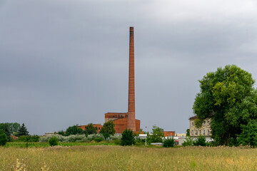 Old brick factory with a tall chimney surrounded by greenery and fields, under a cloudy sky.