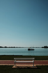A white bench faces Lake Palic under a clear blue sky in Serbia. Calm water, minimalism, and...